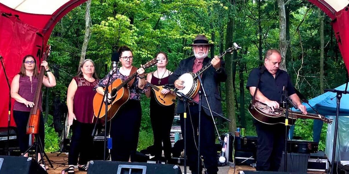 6 musicians on stage playing instruments with trees in the background