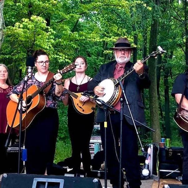 6 musicians on stage playing instruments with trees in the background
